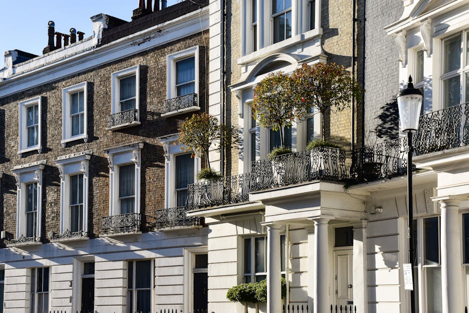 A row of elegant white terraced houses with ornate architectural details, large windows, and decorative balconies located on a busy urban street during daytime. In front of the buildings, several cars