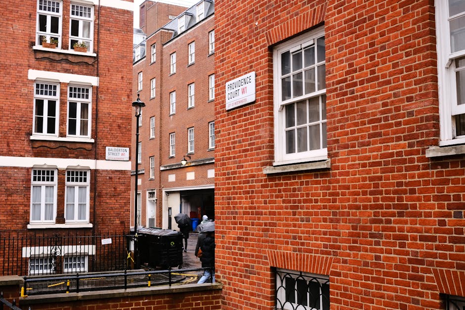 Photograph taken from a narrow street in Marylebone, showing the exterior of a red brick residential building with white-framed sash windows. Two individuals are visible near the entrance of a building, carrying large boxes wrapped in plastic or fabric, engaged in what appears to be the process of home relocation or furniture transport. The boxes are arranged on the pavement close to a black wheelie bin, with a trolley and straps possibly used for moving heavy items. The scene captures the typical urban environment with multi-storey buildings, street signs indicating 'Balderton Street W1' and 'Providence Court W1', and a lamppost, all under natural daylight. The setting reflects professional removals in progress, focusing on the logistics of packing and moving within a busy Marylebone street, as managed by Marylebone Movers.