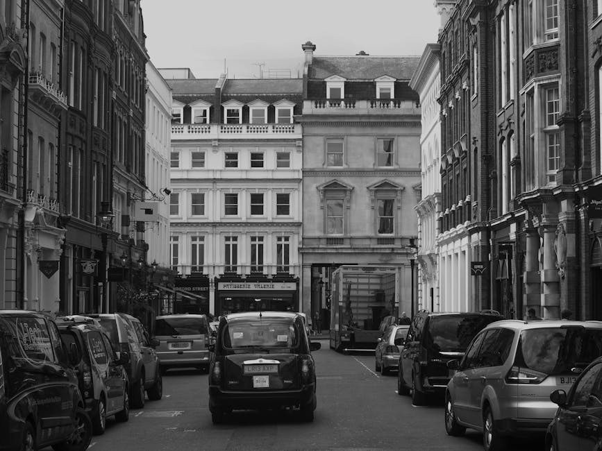 Black and white photograph of a narrow street in Marylebone, London, featuring cars parked along both sides of the road, with some vehicles including a small black car and a larger van visible. In the background, there are classic-style multi-storey buildings with ornate facades, large windows, and sloped roofs. The foreground shows a loading area at the entrance of one of the buildings, where boxes and wrapped furniture might be staged for a home relocation process. The scene suggests the logistical challenge of navigating tight, historic streets for furniture transport and packing during a moving service, with Marylebone Movers potentially involved in the loading process, using blankets and straps to secure items for transportation. The lighting is natural, indicating daytime, and the overall atmosphere emphasizes the detailed environment where careful planning and equipment are necessary to facilitate successful removals on a busy, urban street.